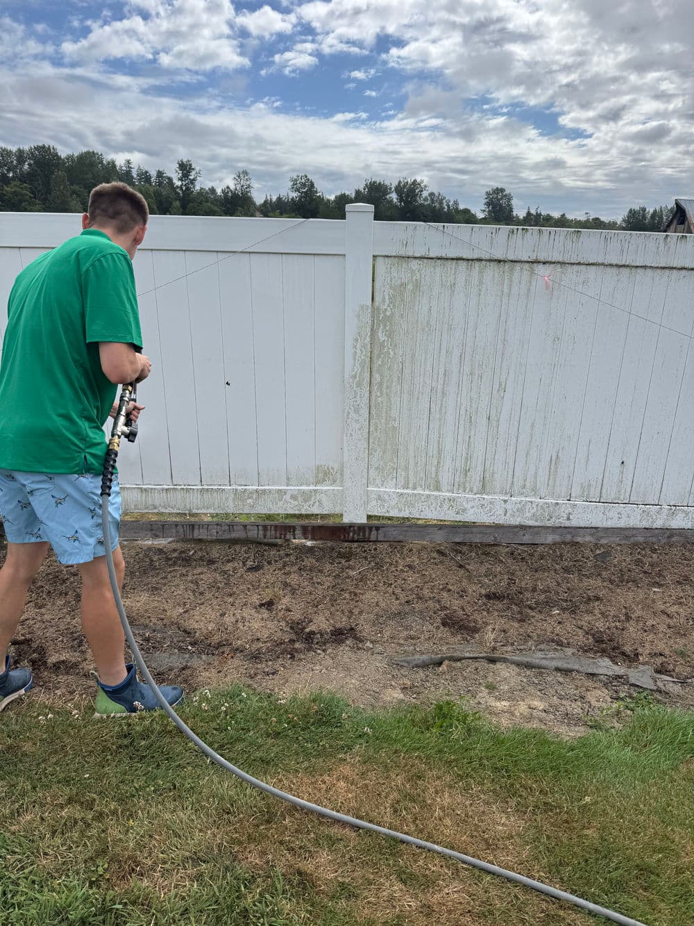 Person pressure washing a dirty white fence in a grassy yard under a cloudy sky.