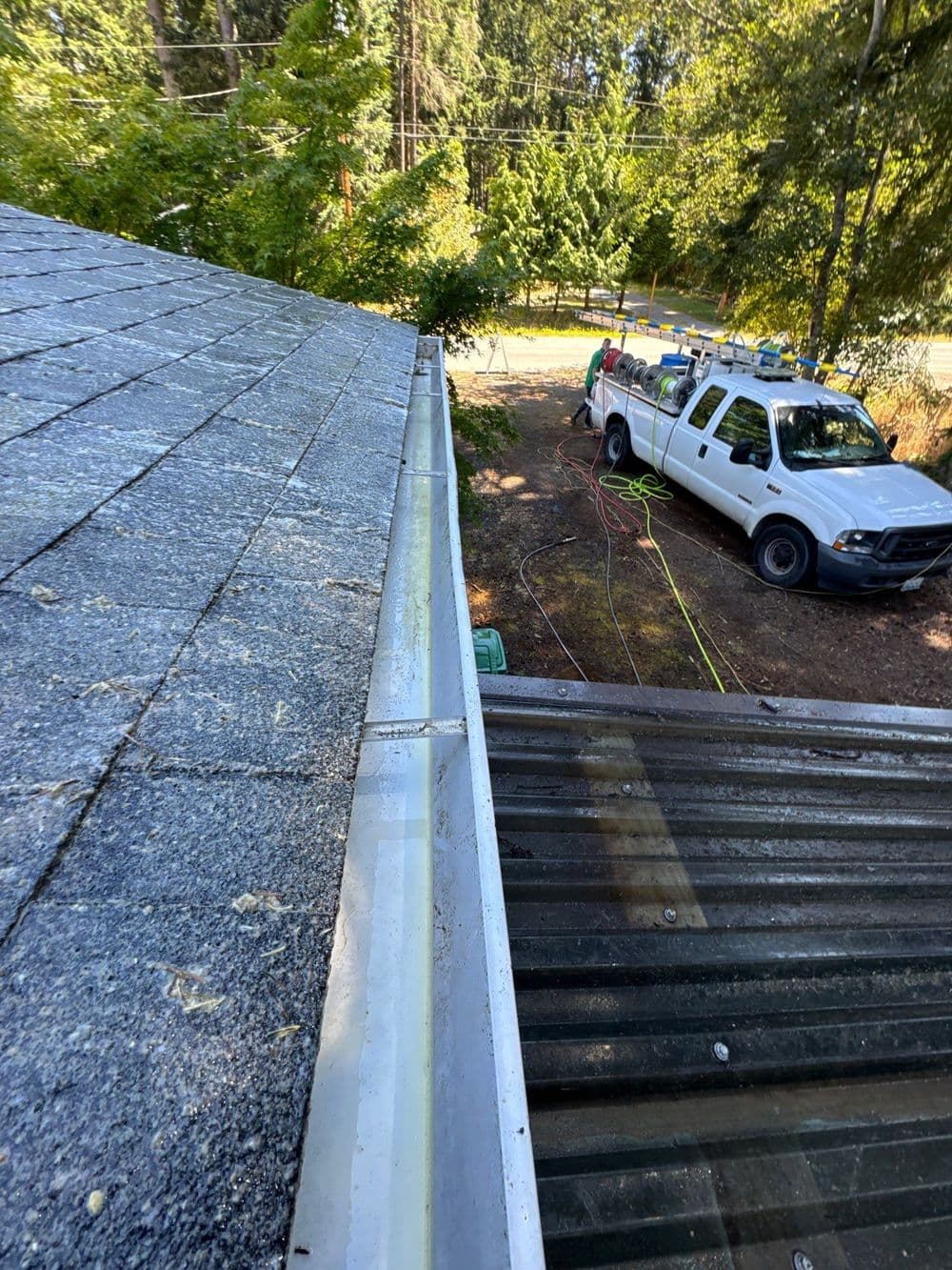 Roof edge with shingles and gutter, service truck in background, surrounded by greenery.