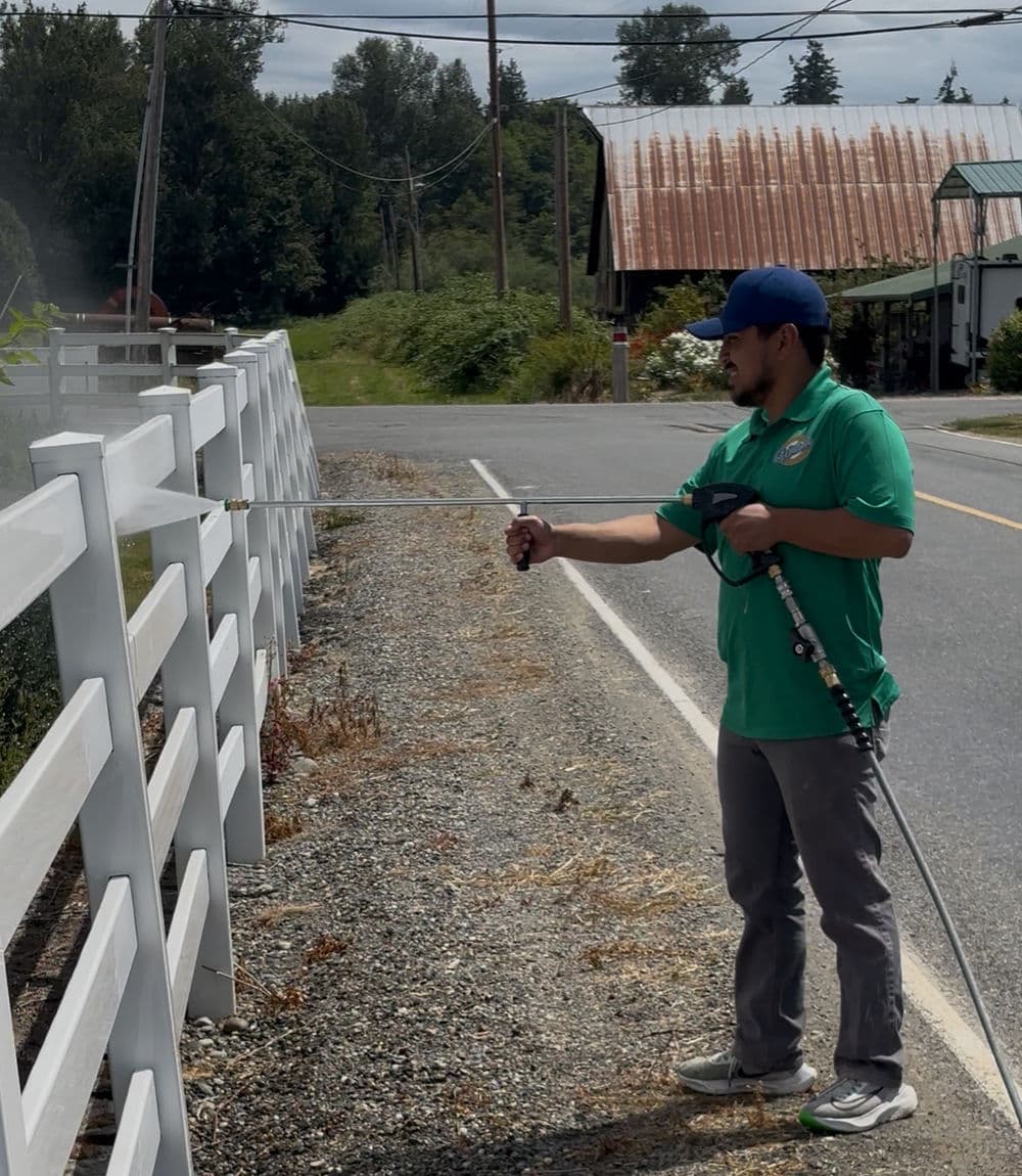 Man cleaning a white fence with a pressure washer along a rural road.