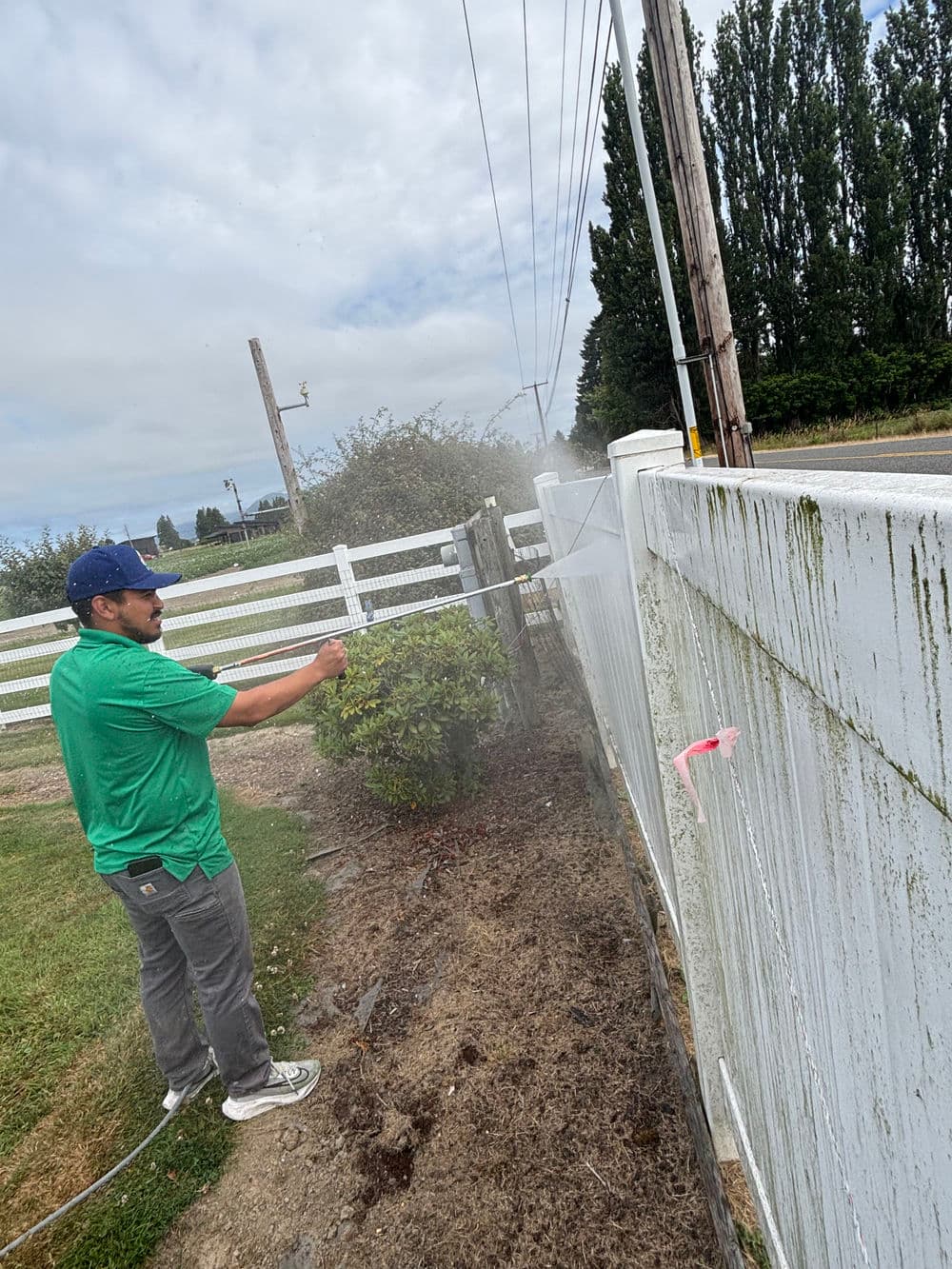 Man power washing a dirty white fence with greenery in the background on an overcast day.
