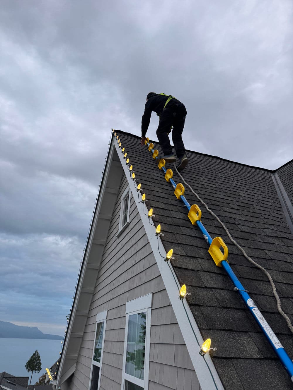 Worker installing Christmas lights on a roof with a ladder under a cloudy sky.