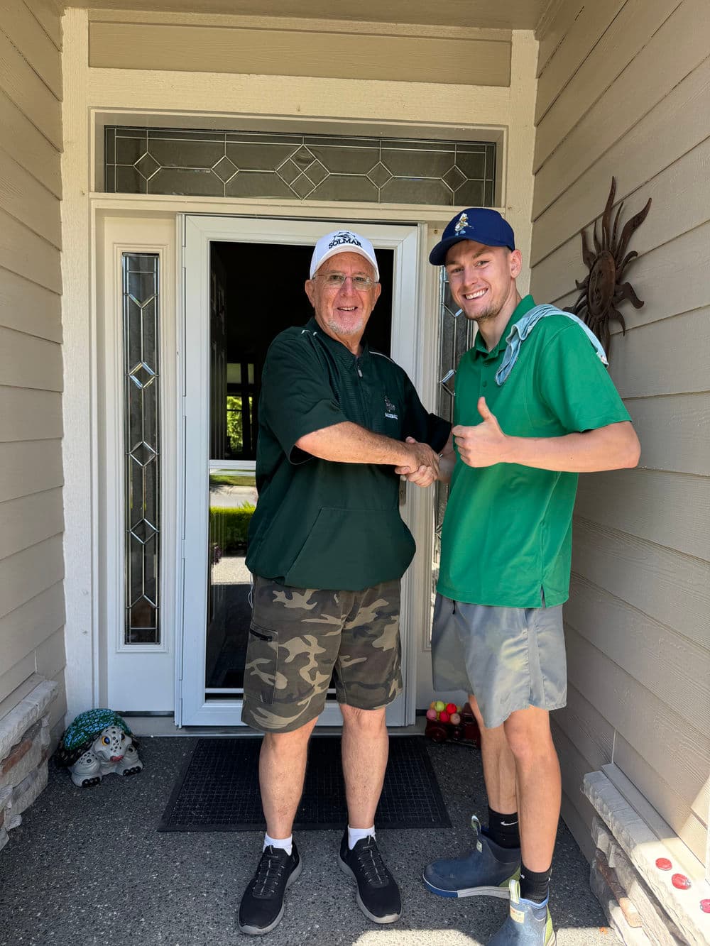 Two men shaking hands at a home entrance, both wearing green shirts and smiling.