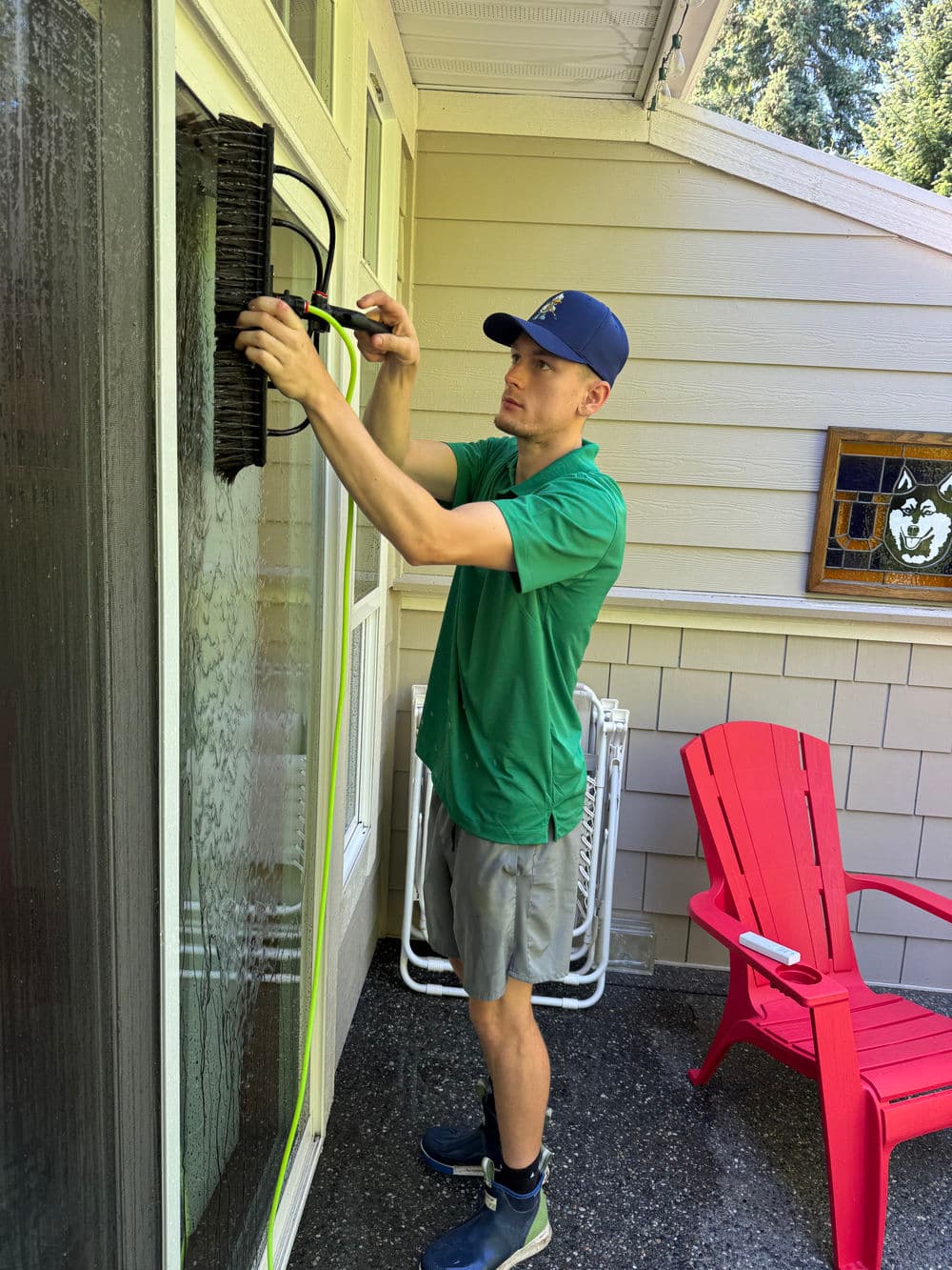 Man cleaning a window screen on a home exterior with a hose and brush. Red chairs in background.