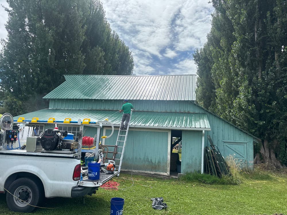 Person cleaning a green barn roof using a ladder, with a truck and tools nearby.