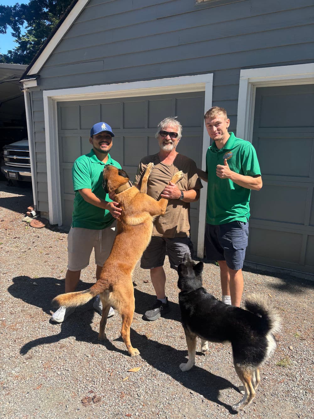 Two smiling men in green shirts with three dogs outdoors by a gray garage on a sunny day.