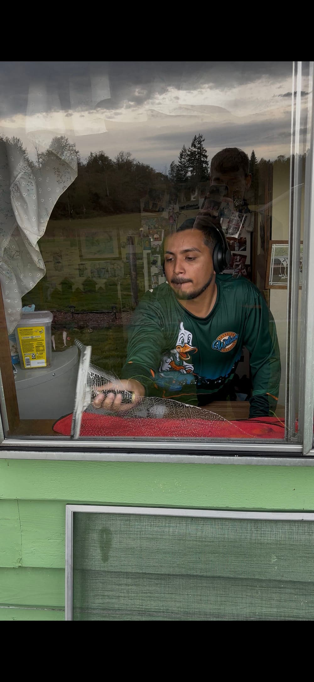 Man cleaning a window indoors, wearing headphones and a green long-sleeve shirt.