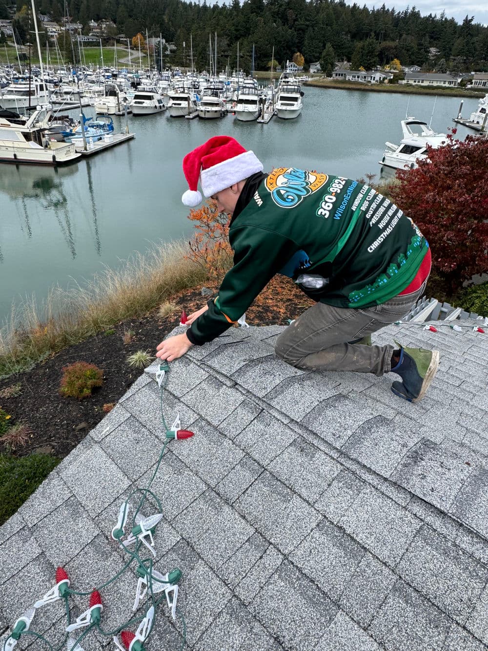 Person wearing a Santa hat installing holiday lights on a roof near a marina.