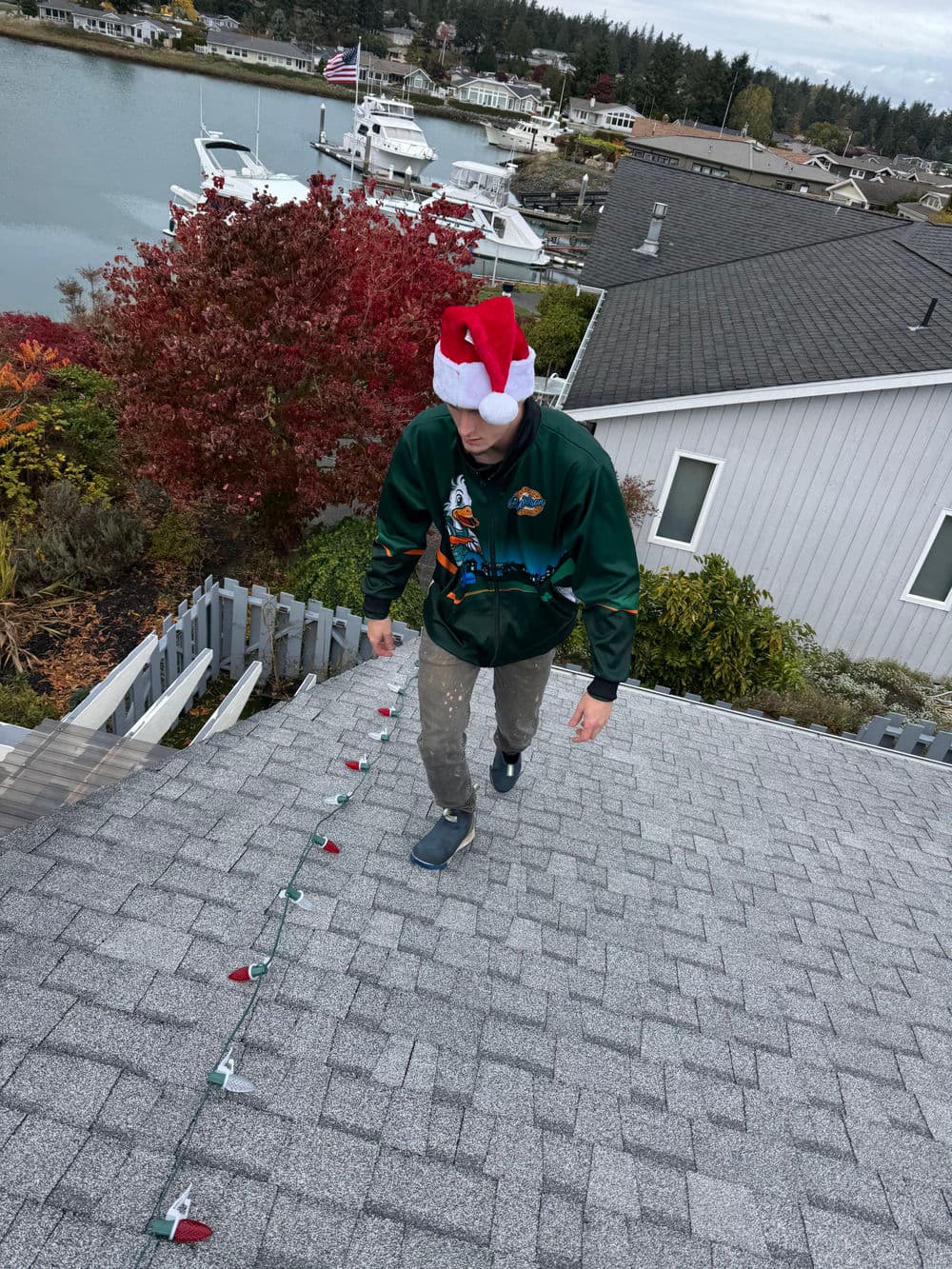 Person in Christmas hat installing holiday lights on a rooftop by a waterfront neighborhood.